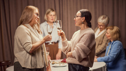 Focus on two women of foreground, standing, talking, discussing news, drinking wine with other senior women, friends on background. Concept of elderly lifestyle, positive emotions