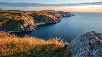 Naklejka premium Scenic coastal view of a tranquil bay with rugged cliffs and golden grass at sunset under a clear sky