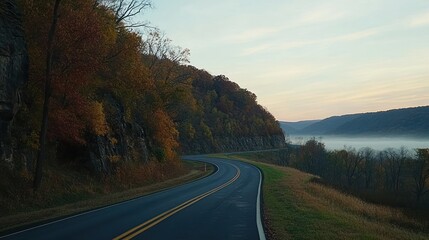 Fototapeta premium Winding road through autumn foliage overlooking misty valley at dawn.
