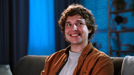 Young man spending evening at home sitting on sofa in living room, watching tv, evening with movie or news program, positive smiling expression, close up.