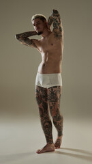 Full-length portrait of confident young young tattooed male model standing with his hands behind head, emphasizing his full-body tattoos and strong physique. Concept of male beauty, fashion, body care