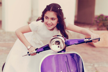 Smiling Girl wearing a first communion dress sitting  on a purple scooter