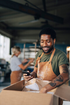 Smiling male baker holding smart phone and checking for supplies in cardboard box while working at bakery