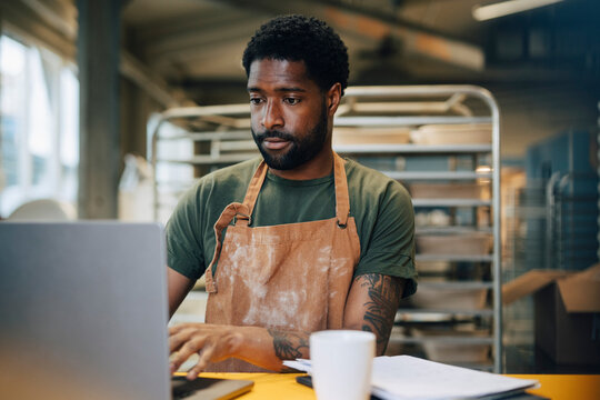 Male baker wearing apron and working on laptop at bakery