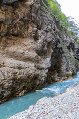 Steep cliffs hang over mountain river with emerald-clear water. Stormy Chegem River flows in deep crevice along bottom of Chegem Gorge. In depths of gorge, semi-darkness reigns.