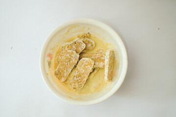 Raw tempeh that has been cut in a white bowl containing dough, with a white background.