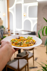A hands holds a plate of vibrant pasta garnished with fresh herbs. The background features a modern dining space with soft lighting and contemporary decor, creating an inviting atmosphere.