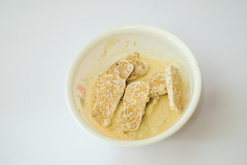 Raw tempeh that has been cut in a white bowl containing dough, with a white background.