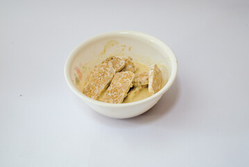 Raw tempeh that has been cut in a white bowl containing dough, with a white background.
