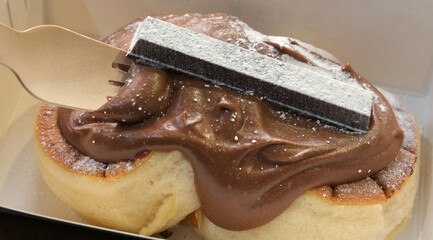 Close-up of a fluffy pancake topped with rich chocolate cream, powdered sugar, and a chocolate bar garnish, served in a takeout box.
