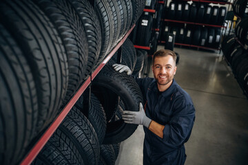 In professional uniform near the shelve. Man worker is maintenance station with tires, wheels