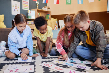 Group of young schoolkids playing alphabet game on floor and laying cards while learning English together