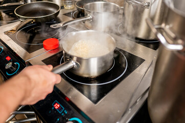 A cook stirs a pot of steaming rice on an induction stovetop. Various pots and pans surround the...