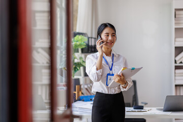 Young business asian woman standing holding documents and talking on smartphone in workplace office
