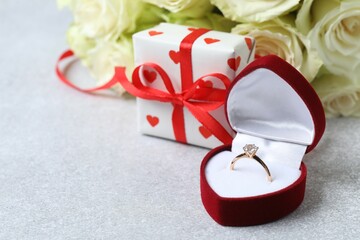 Beautiful shiny gold wedding ring with big diamond gemstone in rich red velvet box with gift box and  white roses on grey background
