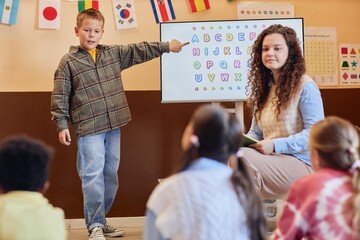 Full length portrait of young boy answering question standing in front of class and pointing at...