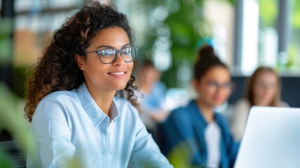 Fototapeta premium A woman with curly hair smiles while working on a laptop in a bright, green environment, surrounded by colleagues.