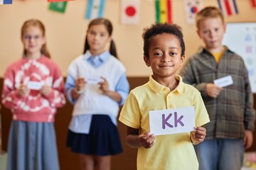 Waist up portrait of African American boy holding letter card and looking at camera with group of children learning English in background