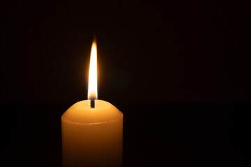 A single burning candle flame or light glowing on a big white candle isolated on black or dark...