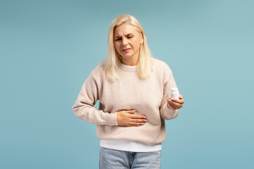 Mature woman suffering from stomach ache holding pill bottle on blue background