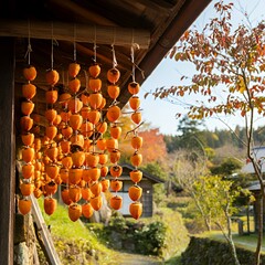 A traditional Japanese scene of hoshigaki (dried persimmons) hanging under a wooden house&rsquo;s eaves, surrounded by rural autumn landscapes, glowing in warm sunlight.

