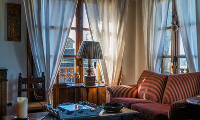 A warm room with sunlight filtering through white curtains, a striped sofa, wooden furniture, a decorative tray, and a candle.