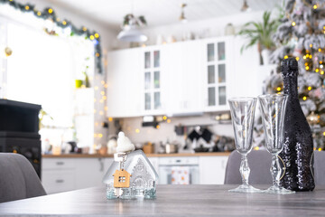 Ice transparent house with a key on table of cozy white kitchen with Christmas tree and Christmas decor with garland lights. Housing, interior, renovation, mortgage, rent in New Year