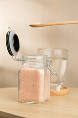 This image depicts a wooden spoon scooping Himalayan salt from a small glass jar to be mixed with water, positioned on a wooden table.