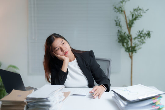 Young Asian businesswoman is feeling bored and tired while working with a laptop and many documents on desk at office, feeling no motivation and lazy, burnout and sleepy