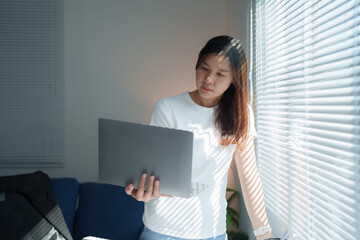 Young Asian freelancer woman working from home, using a laptop computer by the window in her living room, enjoying natural light and a relaxed atmosphere during the day