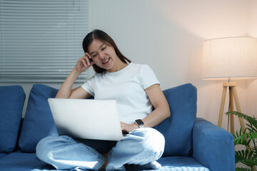 Young asian woman using laptop computer working from home sitting on blue sofa in living room, studying online, watching webinar, making video call, freelancer writing blog