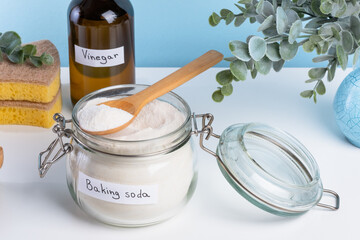 A jar filled with baking soda and a wooden spoon rests on a countertop alongside a bottle of vinegar and cleaning sponges. DIY cleaning method using natural products to remove stains. top view