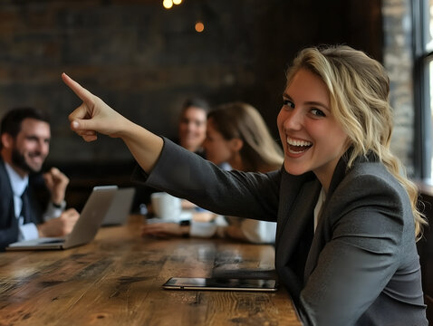 A cheerful businesswoman in a suit points playfully during a team meeting in a modern, rustic office setting.