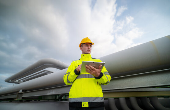 Worker Inspects Industrial Pipelines While Using a Tablet in a Safety Vest Under Cloudy Skies