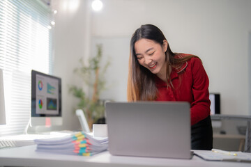Naklejka premium Asian businesswoman is standing at her desk, laughing while working on her laptop, with a pile of paperwork and a desktop computer in the background