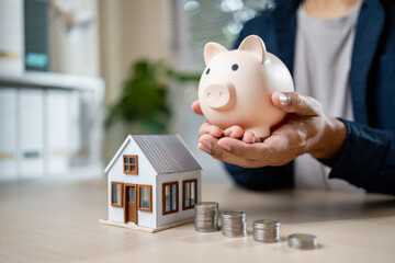 Person holding piggy bank near house model and coins, symbolizing savings