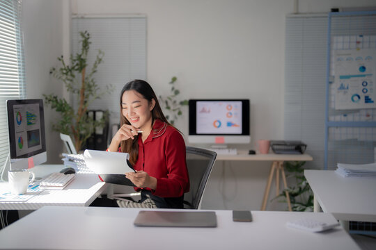 Young asian businesswoman holding pen and reading financial documents, smiling and working at office desk with computer showing graphs, startup business concept