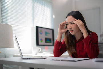 Young office worker suffering from a migraine, experiencing a strong headache, massaging her temples and feeling tired while working with laptop and computer in office