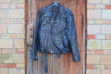 Fototapeta premium one leather old worn dirty biker classic unbuttoned with iron zippers stylish fashion mens black with belt and buckle jacket hanging on wooden brown doors near brick wall