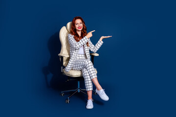 Fototapeta premium Confident young woman with red hair in trendy white checkered suit sitting on chair against dark blue background.