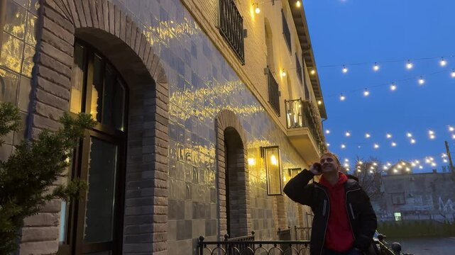 Man having a pleasant phone conversation outside a building decorated with string lights at dusk