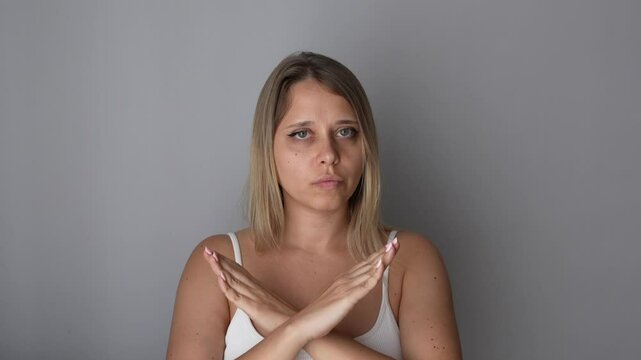 Young blonde woman showing a sign of denial, refusal, rejection crossing her arms in front of her on a grey background. Girl making a gesture by turning her head left and right, giving negative answer