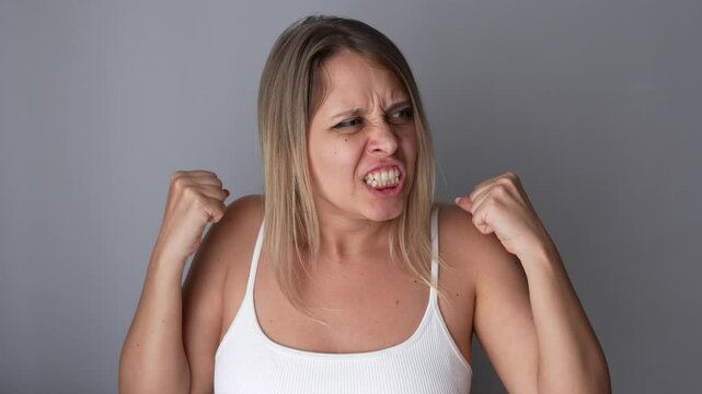 Angry furious young caucasian blonde woman clenching her fists punching with her hands on a dark grey background. Negative emotions, bad mood. Enraged girl feels rage