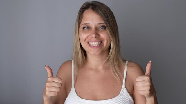 A young caucasian happy excited smiling blonde woman showing thumb up like gesture with two hands nodding her head in approval on a dark grey background. Consent, agreement