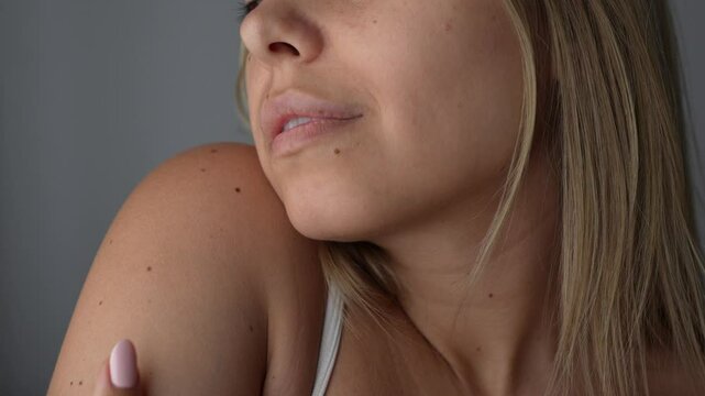 Young attractive caucasian blonde woman stroking her shoulder with hand enjoying the softness of her smooth skin and hugging herself on a grey background. Love of self, skin care. Close up