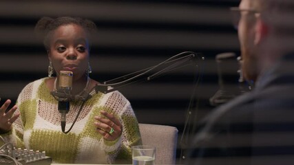 Over-the shoulder shot of young African American woman wearing shiny earrings speaking in mic while talking to man in eyeglasses seen from partition wall of modern broadcasting studio