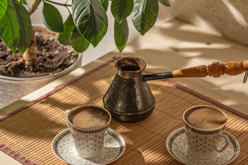 Two cups of coffee and cezve on the table in the patio lit by morning sun.	