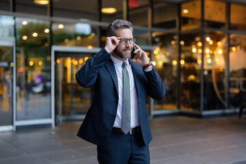 Mature Hispanic businessman in suit and glasses having business call and talking on phone outdoor