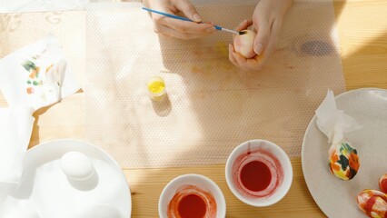 Close-Up of a Woman Adding Yellow Paint to an Easter Egg, with Dye Bowls on the Table.