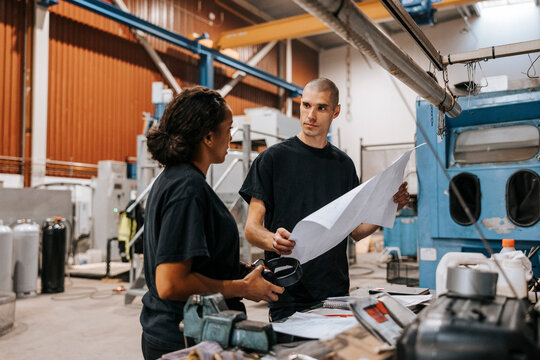 Mature male worker holding blueprint while discussing with female colleague in manufacturing warehouse